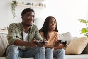 Happy couple playing video games on a cozy couch with wireless controllers—perfect scene for promoting where to buy gaming consoles online.