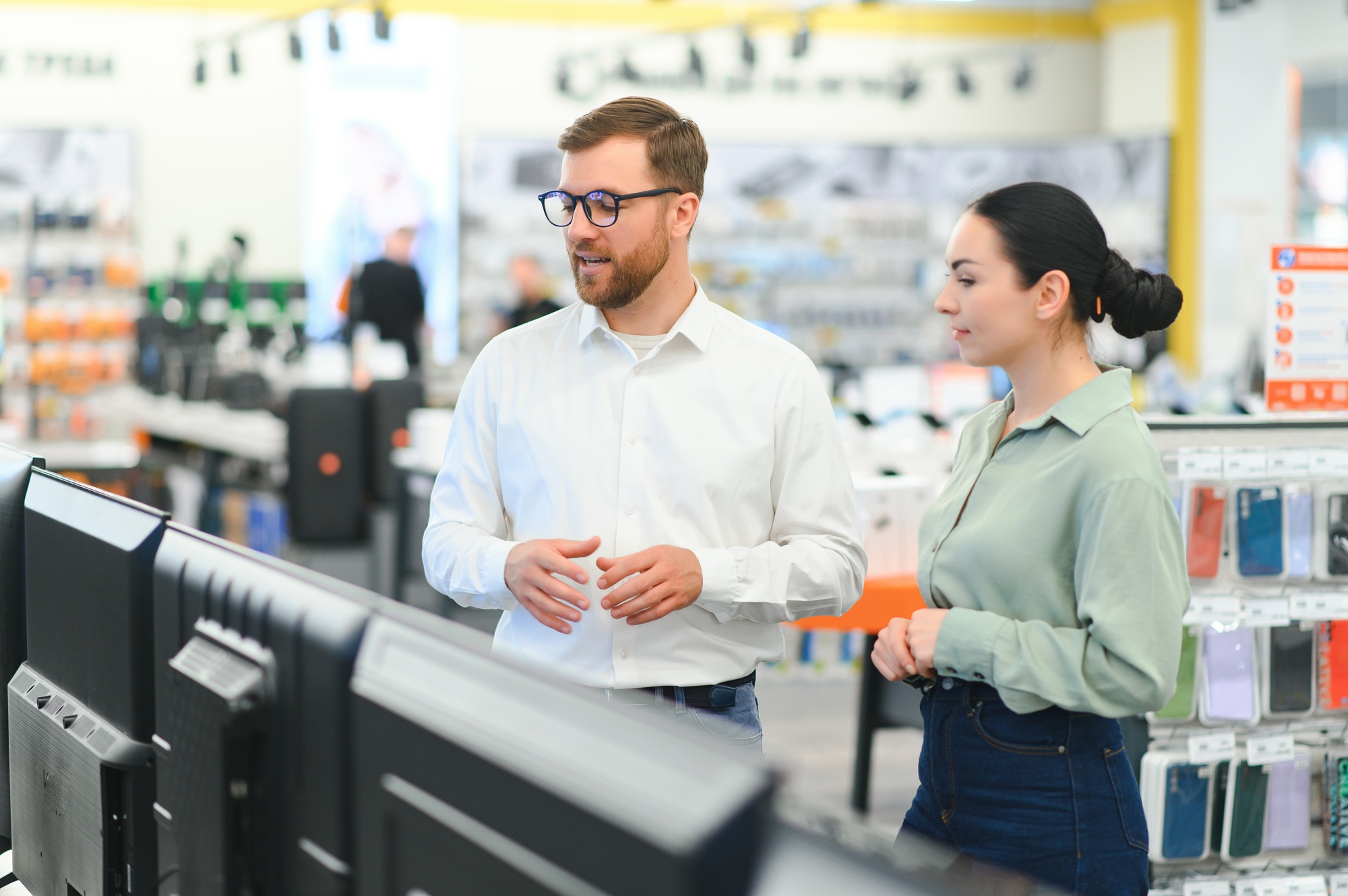 Two customers consulting in front of a TV display section inside a modern electronics retail showroom — perfect brand representation for tech store near me Florida searches and in-store assistance services.

